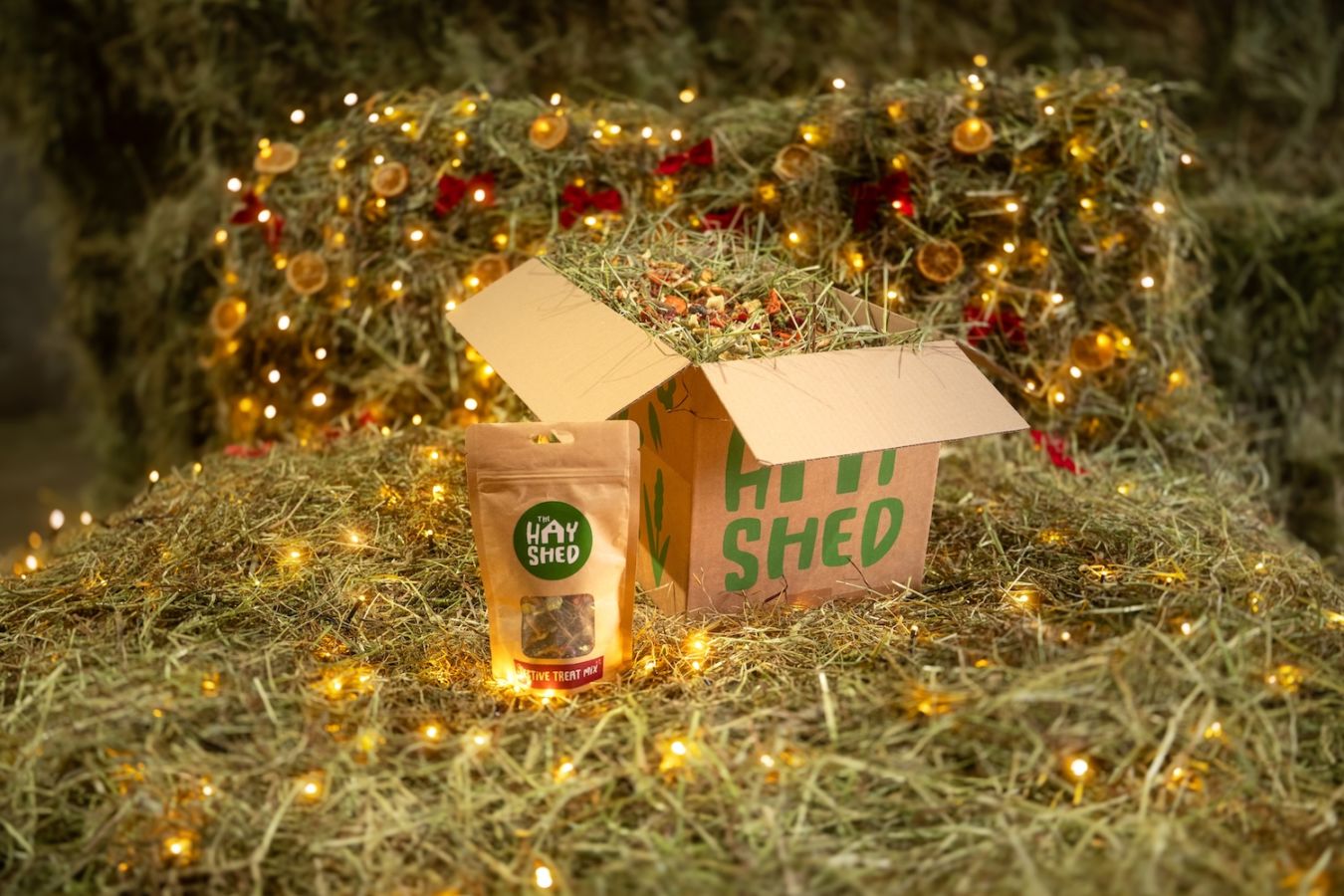 A bag of Festive Treat Mix, a box of hay with a delicious topper sitting on hay with a Christmas background at The Hay Shed